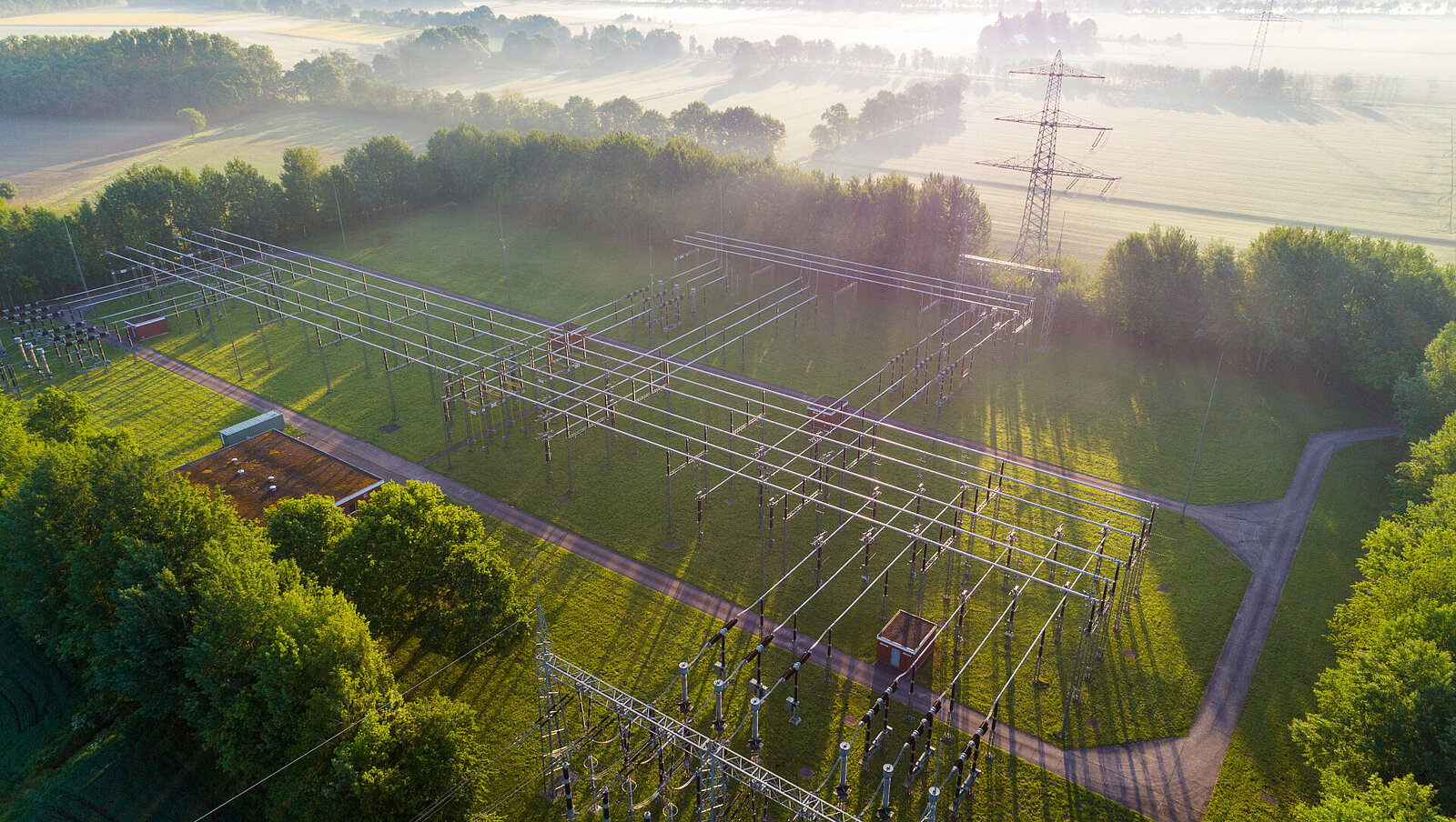 Luftaufnahme eines Umspannwerks, umgeben von grünen Bäumen und Feldern, mit Stromleitungen und Strommasten, die sich im frühen Morgenlicht durch die Landschaft erstrecken.