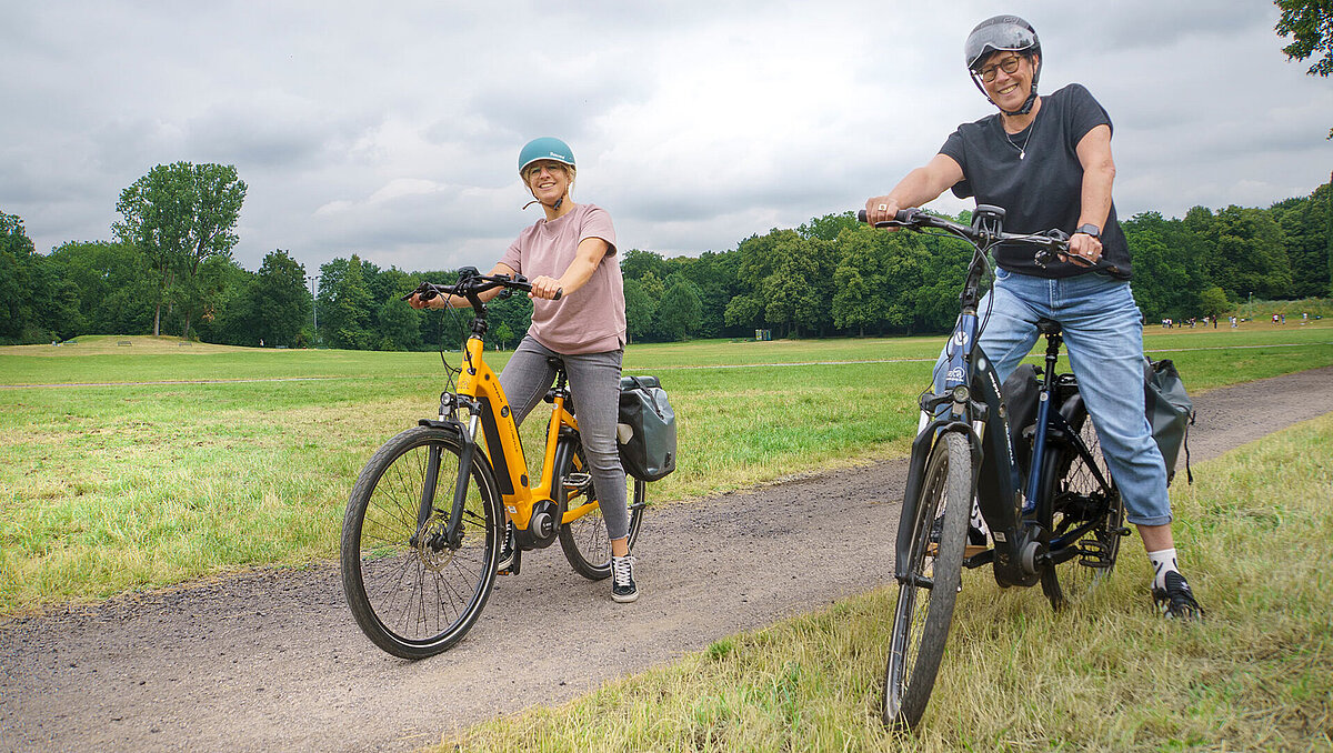 Bild von einem Weg in einem Park. Ein blaues und ein gelbes Pedelec mit jeweils einer Fahrradfahrerin mit Helmen in sommerlicher Kleidung. 