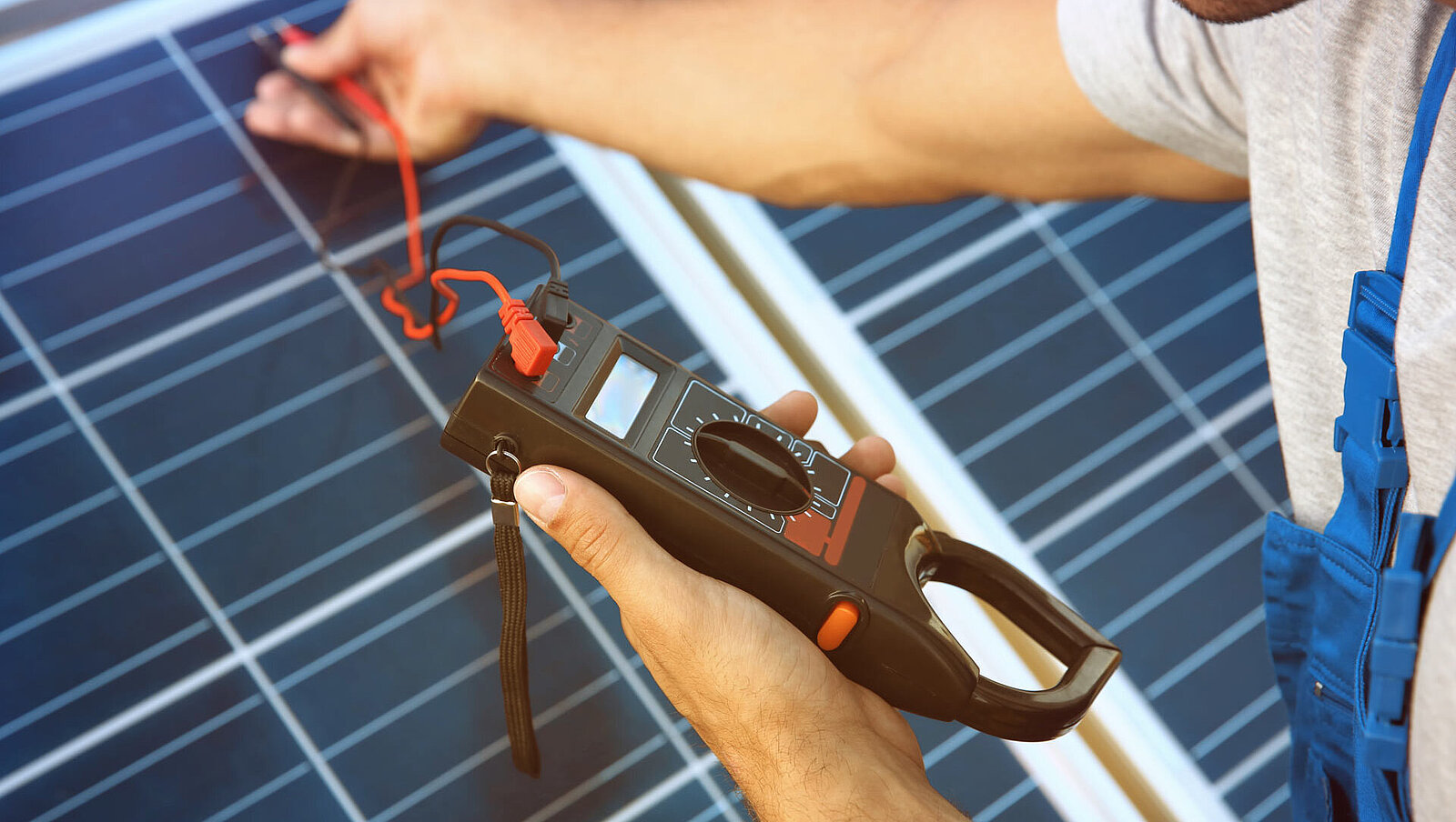 Eine Person in einem grauen Hemd und einem blauen Overall misst mit einem Digitalmultimeter die Spannung an einem Solarpanel.