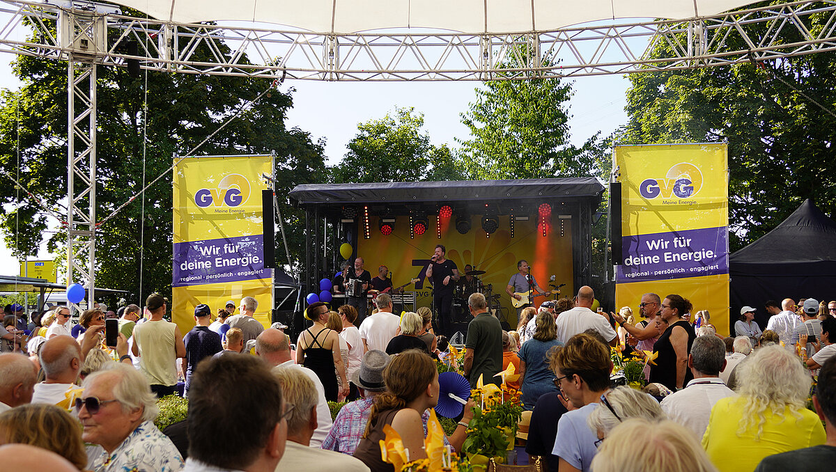 Pubklikum vor einer Bühne die zum Jubiläum geschmückt ist. Eine Band spielt auf der Bühne.