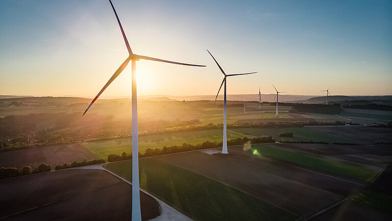 Bei Sonnenuntergang stehen Windräder in einer Reihe auf grünem und braunem Ackerland. Das Sonnenlicht scheint durch die Rotorblätter und die Felder erstrecken sich bis in die Ferne.